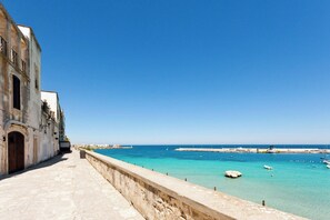 Un paseo marítimo con vistas despejadas al mar y a los barcos, que lleva a un muelle con un muro de piedra en un lado y un edificio con puerta y ventanas en el otro.