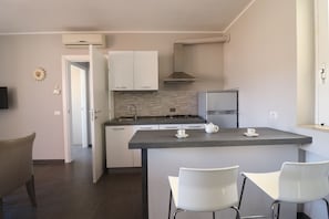 A modern kitchen with white cabinets, a gas stove, and a stainless steel refrigerator. There is a small island with two white chairs and a teapot on it.