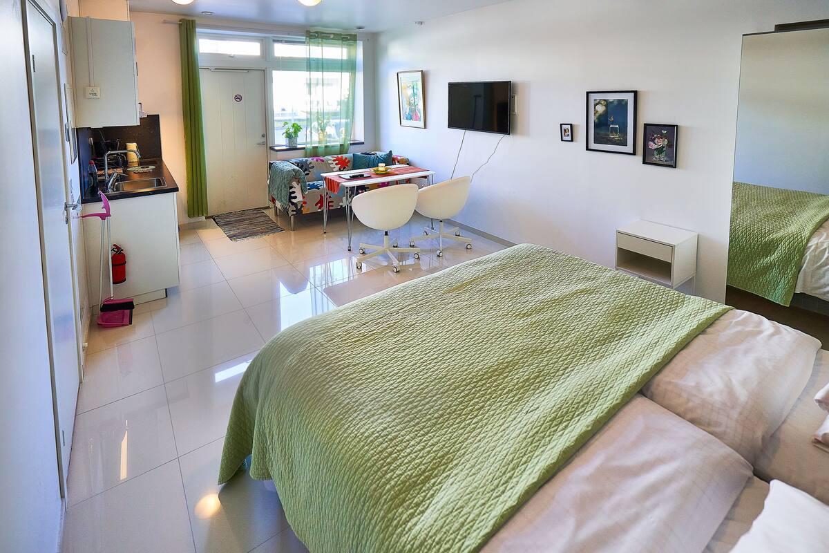 A bedroom with a bed, a desk with chairs, a kitchenette, and a TV on the wall.
