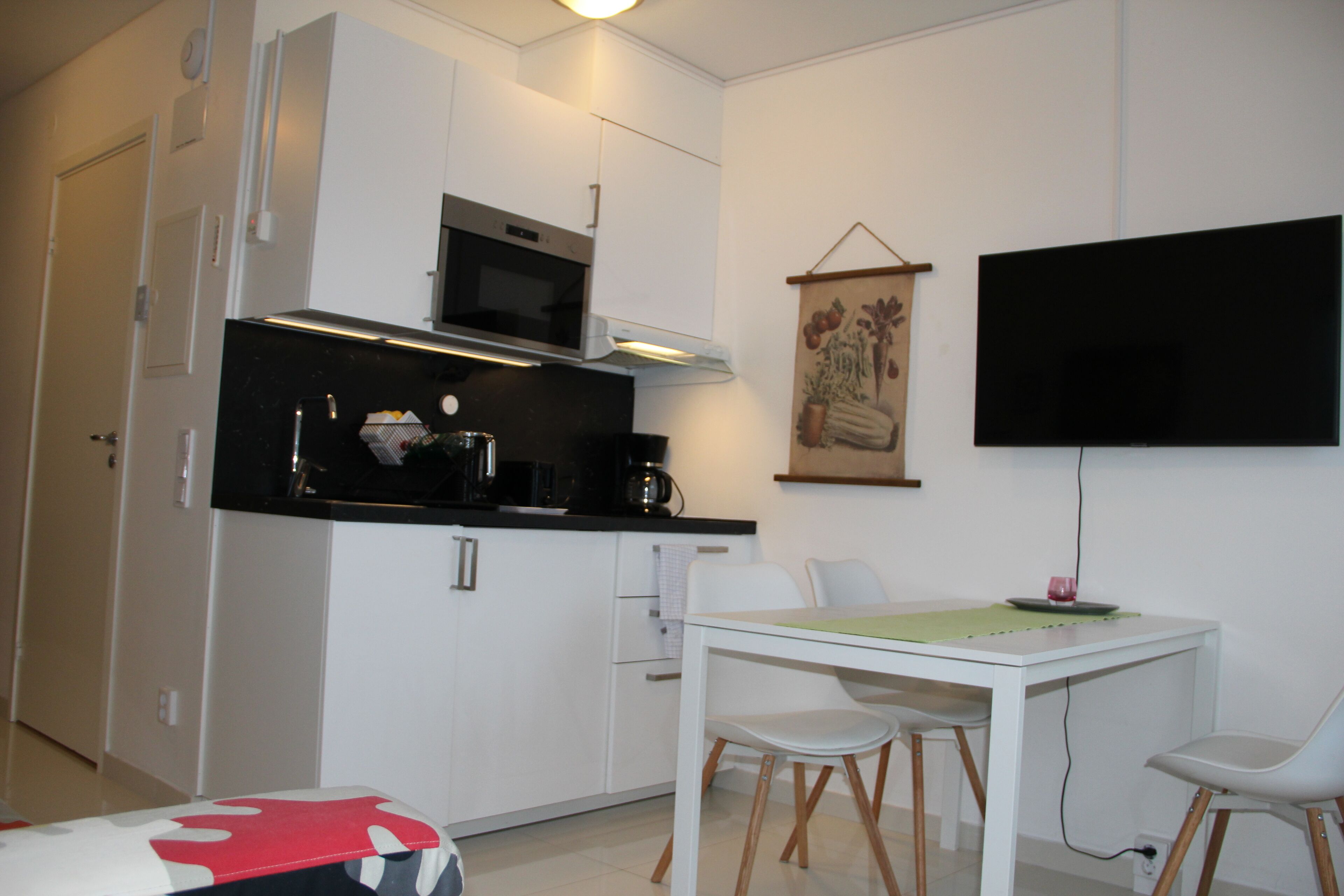 A compact kitchen with white cabinetry, a microwave, and a coffee maker. There is a dining table with chairs and a flat-screen TV mounted on the wall.