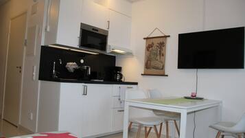 A compact kitchen with white cabinetry, a microwave, and a coffee maker. There is a dining table with chairs and a flat-screen TV mounted on the wall.