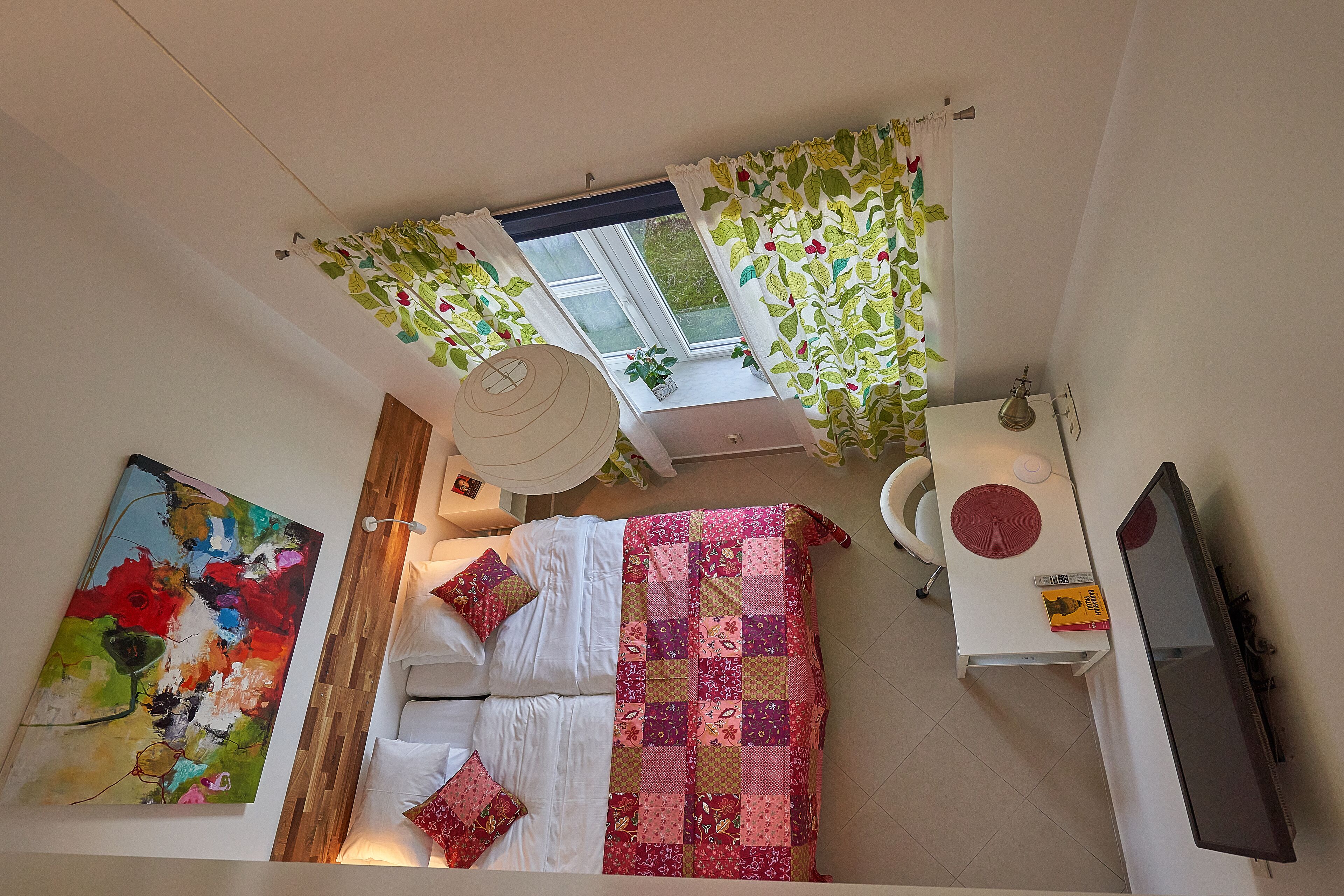 Een slaapkamer met een bed, een sprei, een schilderij, een raam met gordijnen, en een televisie.