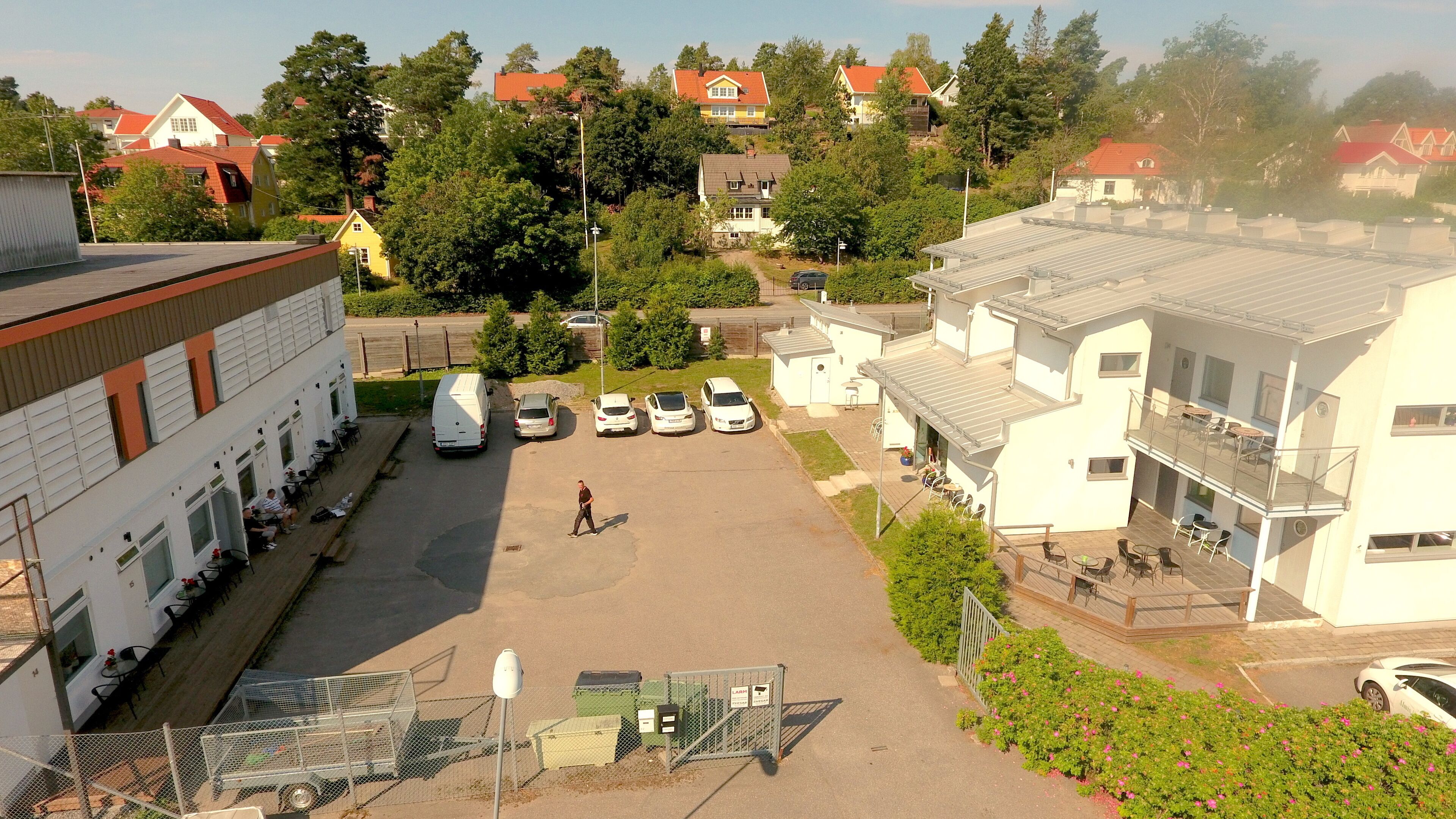 A parking area with several cars and a person walking. There are buildings with balconies and a fenced area with a gate.