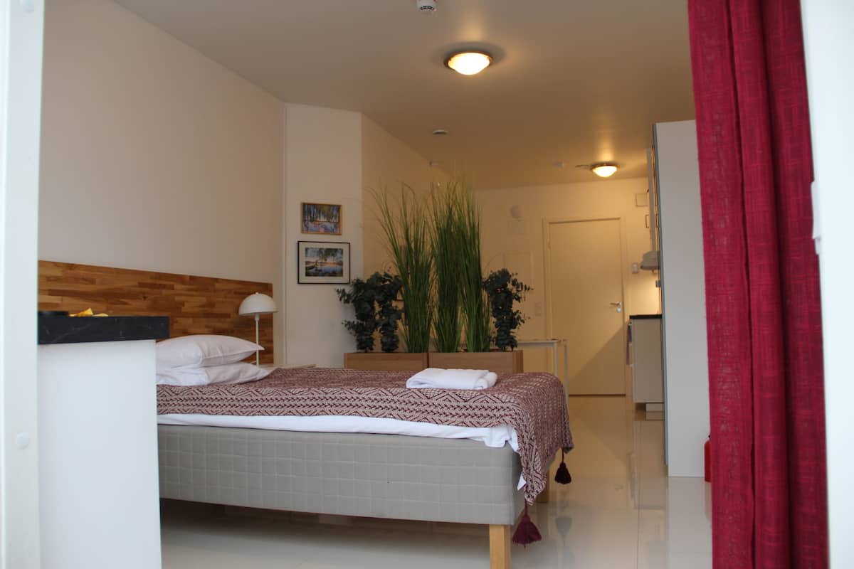 A bedroom with a bed, a nightstand, a lamp, a potted plant, and a door leading to a bathroom.