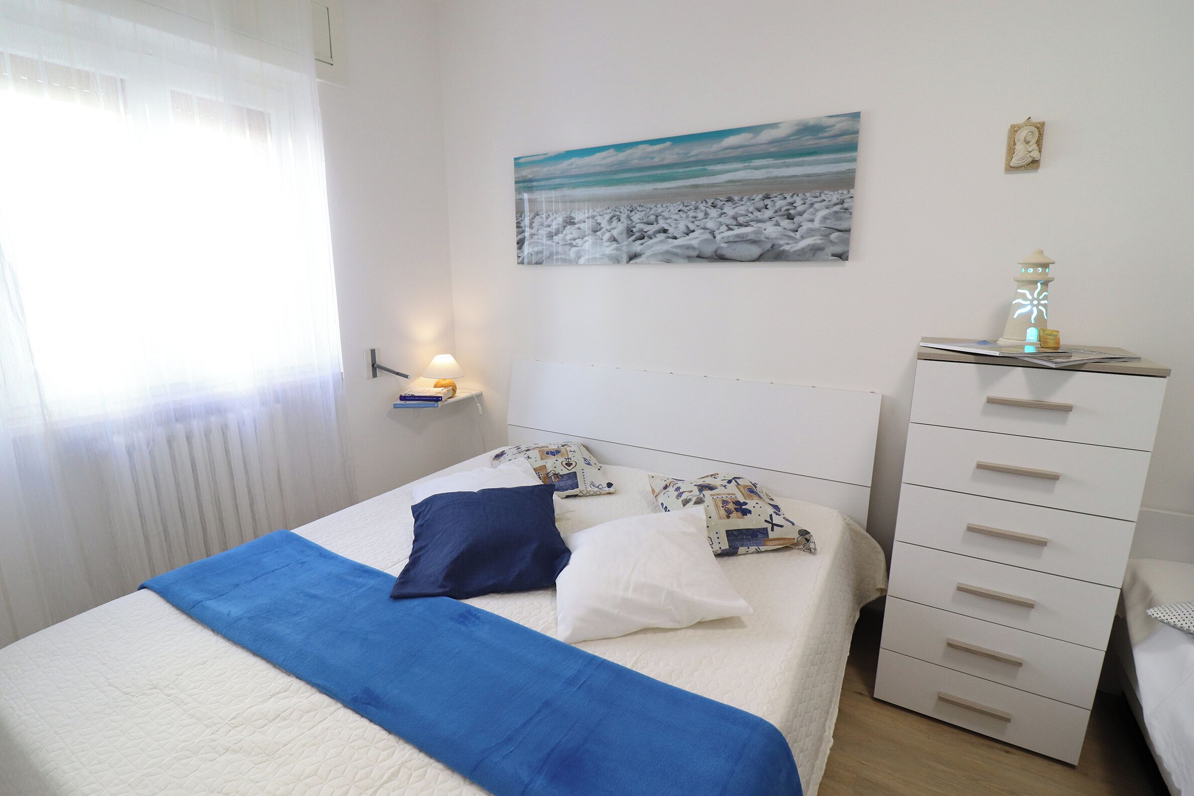 Un lit bien fait, recouvert d’une couvre-lit bleue et blanche, une lampe de chevet et une étagère fixée au mur, sur laquelle se trouvent un livre et un petit objet décoratif.
