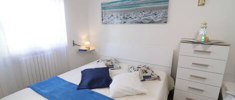 Un lit bien fait, recouvert d’une couvre-lit bleue et blanche, une lampe de chevet et une étagère fixée au mur, sur laquelle se trouvent un livre et un petit objet décoratif.