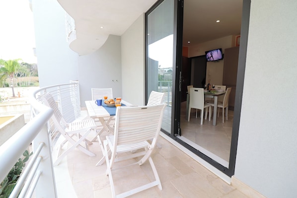 A balcony with white chairs and a table, a TV in the background, and a view of the outside.