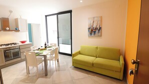 A modern kitchen with white appliances, a dining table with chairs, a yellow sofa, and a painting of sailboats on the wall.