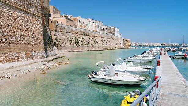 A coastal marina with boats docked along a stone wall and a wooden pier extending into the water.