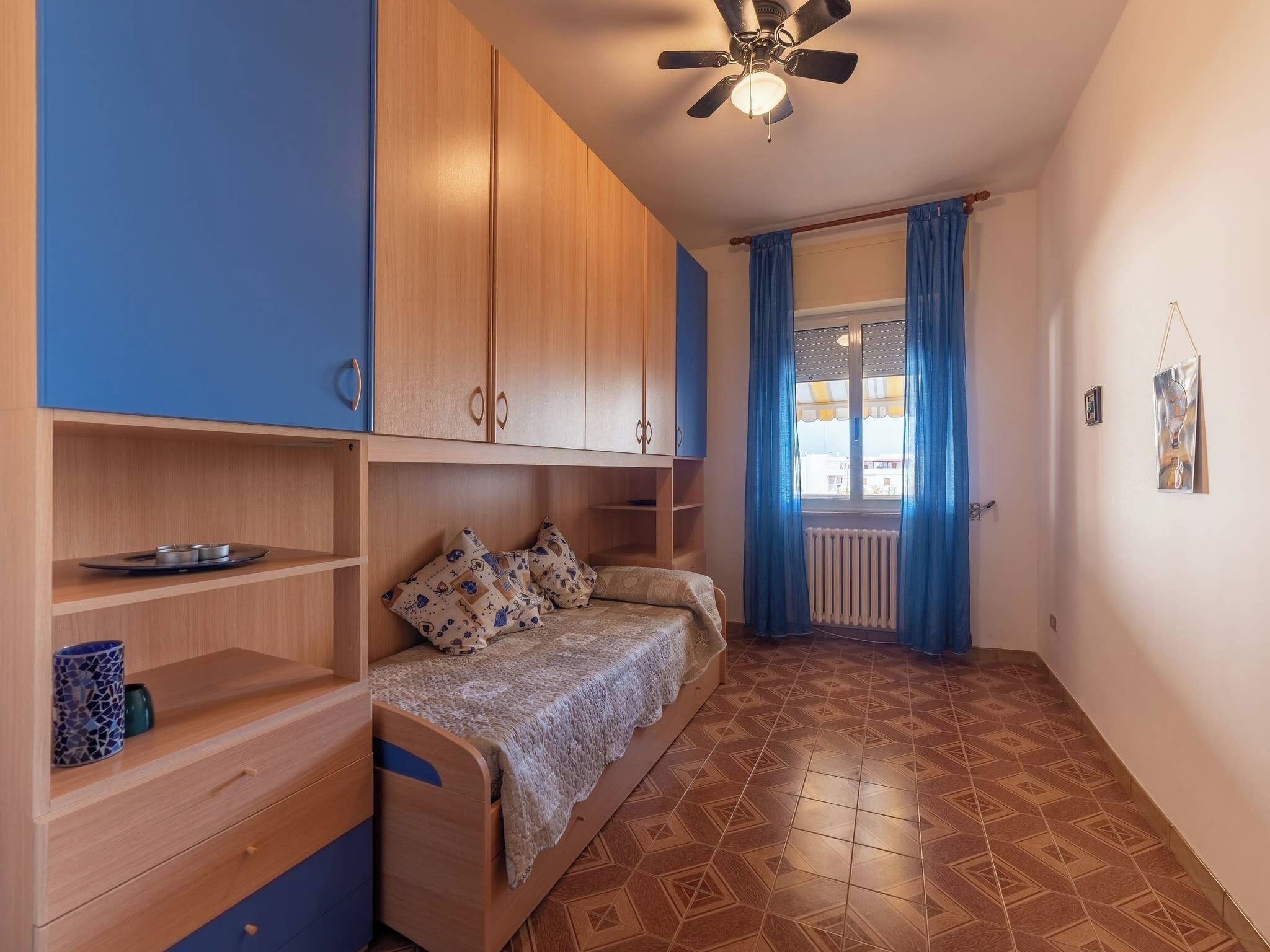 Egy kis méretű hálószoba, amelyben egy ágy, egy fa szekrény és egy mennyezeti ventilátor található.