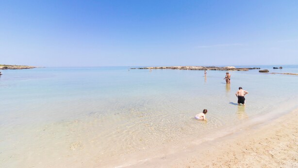 Una playa con aguas transparentes y poco profundas, gente nadando y jugando, y una costa distante.