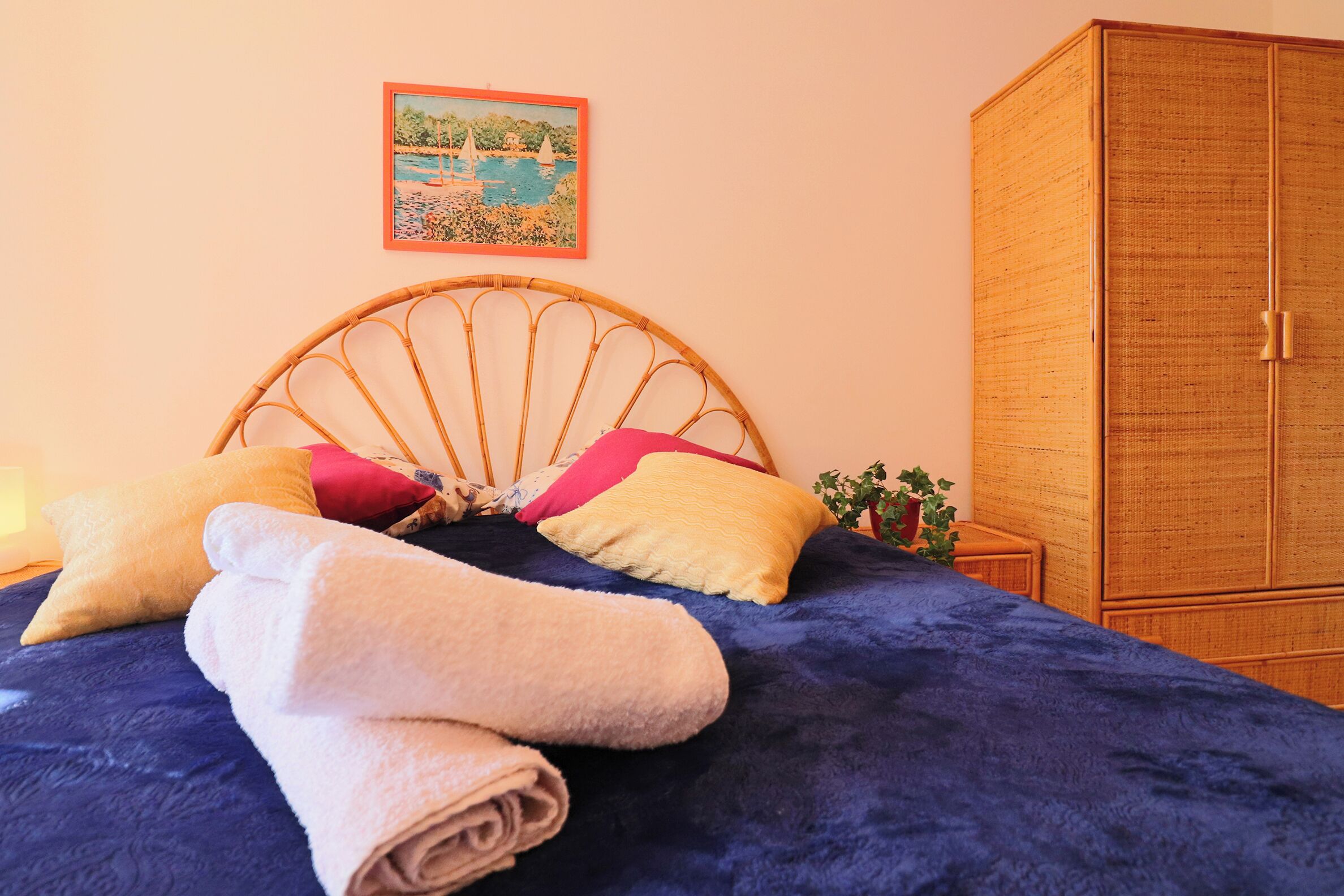 A bed with a blue cover, a wicker headboard, and rolled towels.
