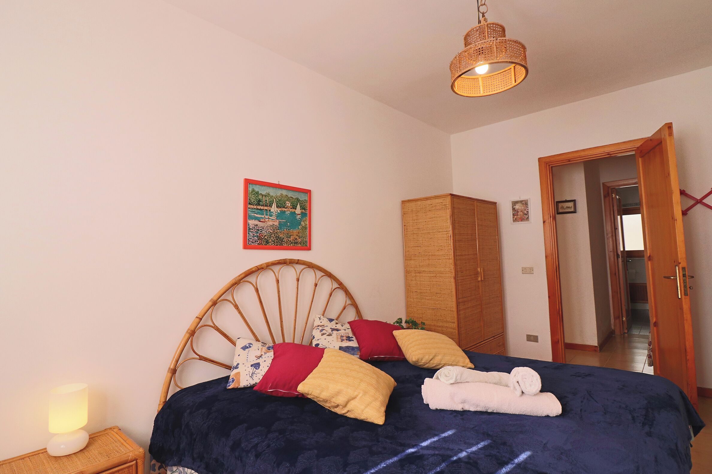 A bedroom with a bed, a nightstand with a lamp, a wooden wardrobe, and a door leading to another room.