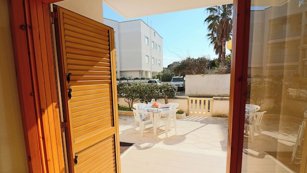 An open wooden door leading to an outdoor patio with a table and chairs, a white building in the background, and a palm tree. - Nice House Cecilia in Otranto (Otranto)