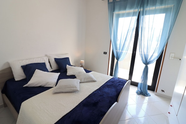A neatly made bed with a white and blue bedspread, a bedside table with a lamp, and sheer blue curtains over a window.