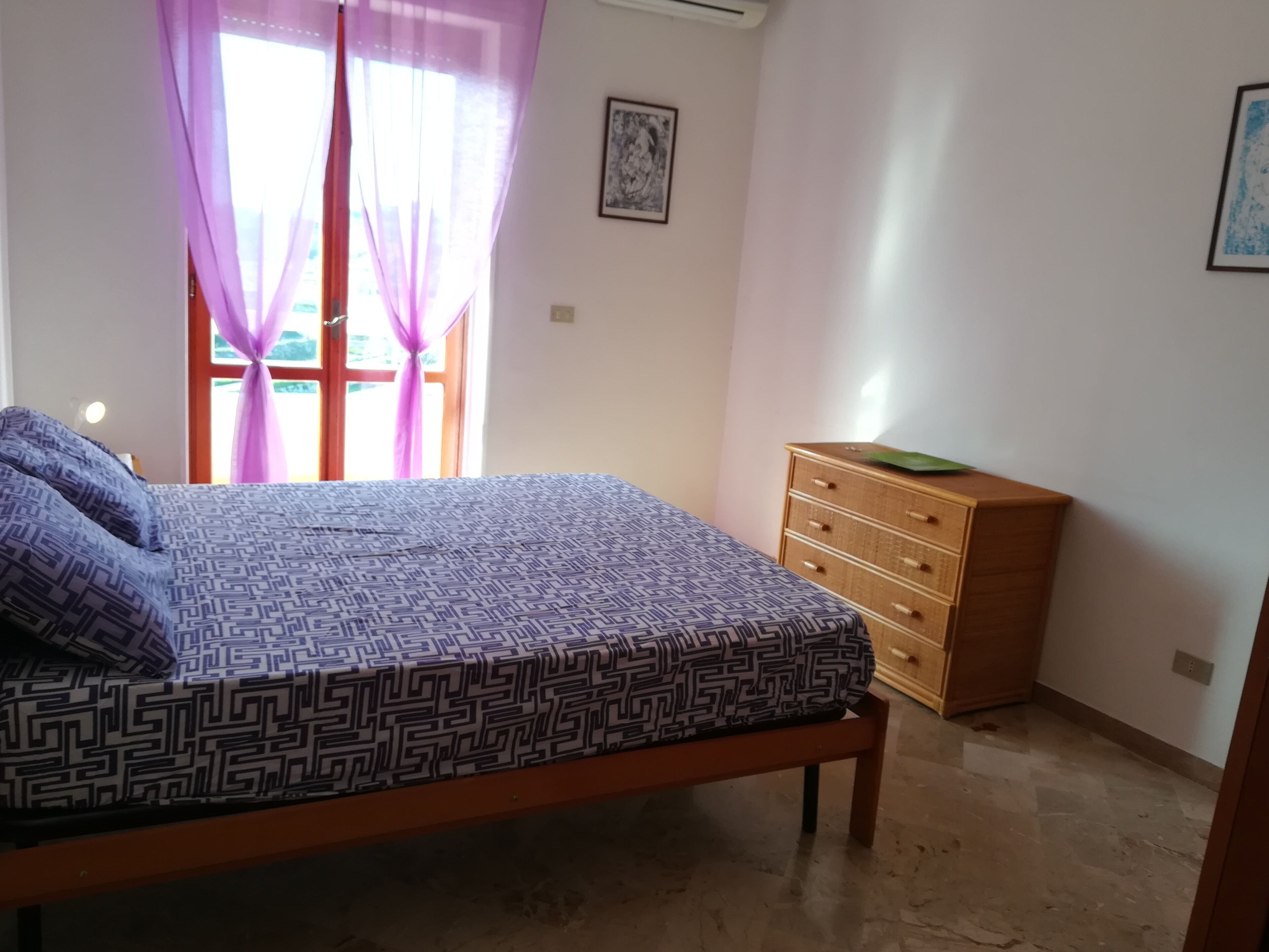 A bedroom with a bed, a wooden dresser, and a window with purple curtains.