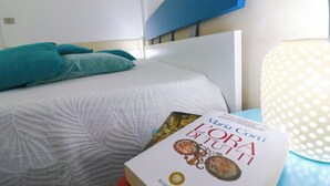 A neatly made bed with a white and teal color scheme, a book titled 'Maria Corti L'ora di Tutti' on the bedside table, and a polka-dotted lamp. - Casa Lucrezia Unique Sea View and Relaxation Near the Historic Centre (Otranto)