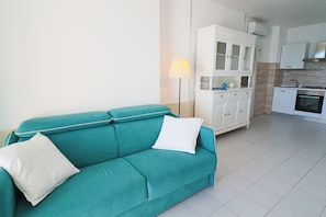 Un sofá color turquesa con dos almohadas, una lámpara blanca, un armario blanco y una cocina con estufa y horno.