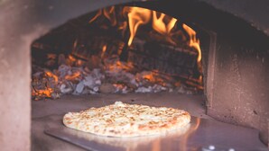 A pizza inside a brick oven with flames visible in the background. - Canvas & Clover (Worcester)