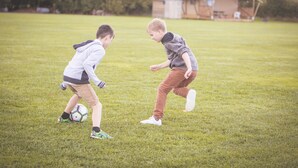 Two children playing soccer on a grassy field with a building in the background. - Canvas & Clover (Worcester)