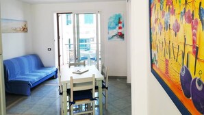 A room with a dining table, chairs, a blue sofa, a painting of a lighthouse, and a view through a glass door. - Holiday Home Barbara (Otranto)