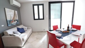A modern living room with a white sofa, a dining table set for two, and a large window with blinds. - Modern Apartment "casa Giuditta Deluxe A Otranto" (Otranto)
