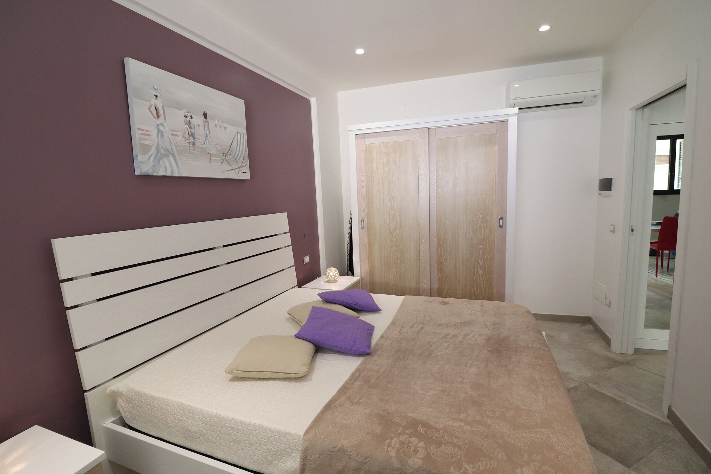 A bedroom with a bed, a nightstand, a painting, a wardrobe, and an air conditioning unit.