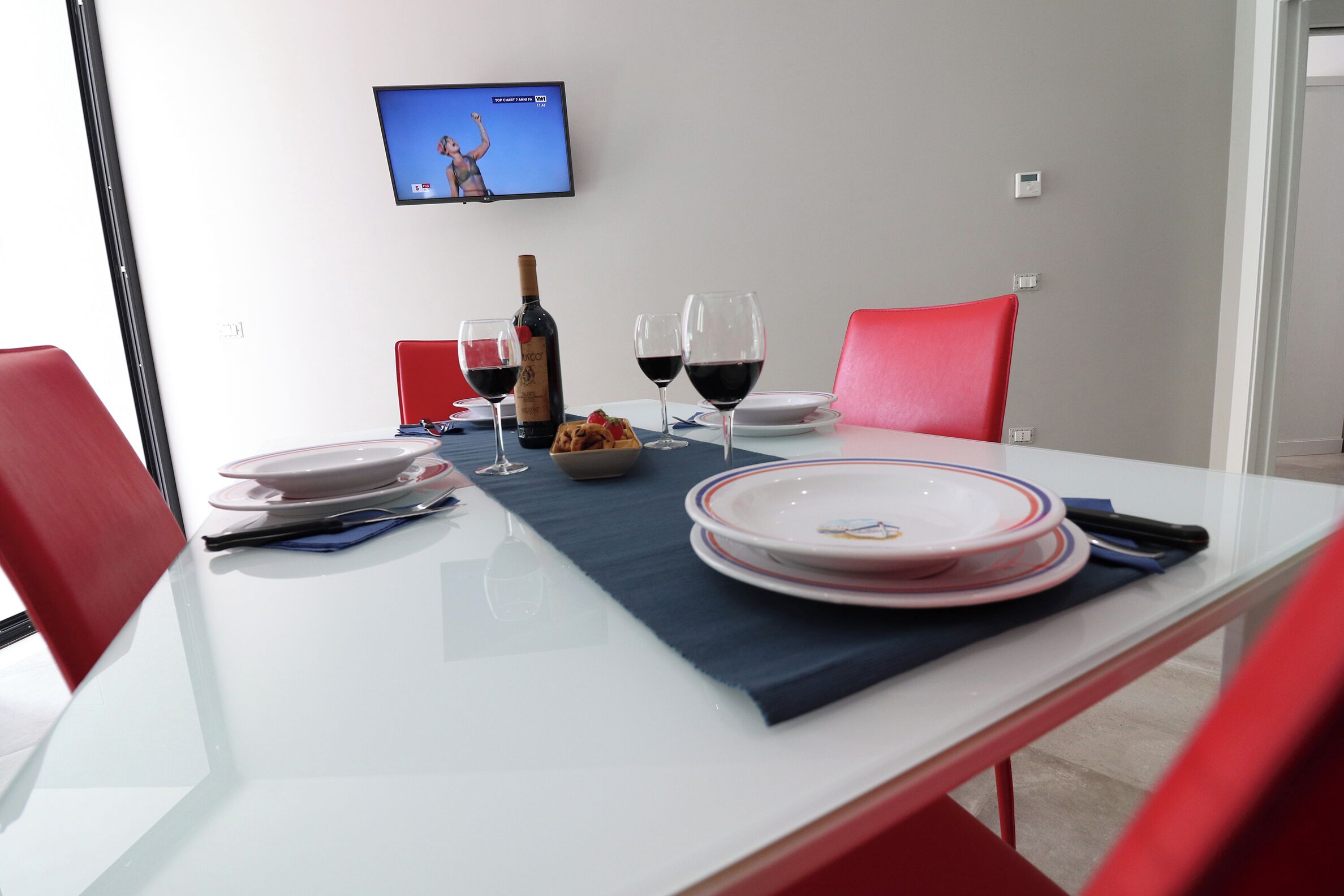 Una mesa de comedor dispuesta con platos, vasos, una botella y un bol, con un televisor montado en la pared al fondo.