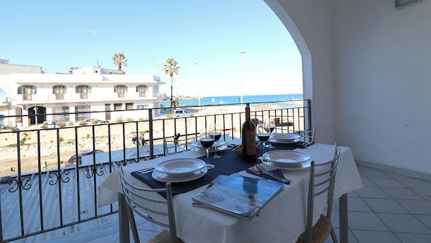 A balcony table set with plates, glasses, and a menu, overlooking a beach with buildings and palm trees. - Casa Vanessa Modern One-bedroom Apartment With sea View in Central Otranto (Otranto)