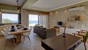 En moderne stue med sofa, spisebord og fjernsyn. Der er en stenvæg og havudsigt.