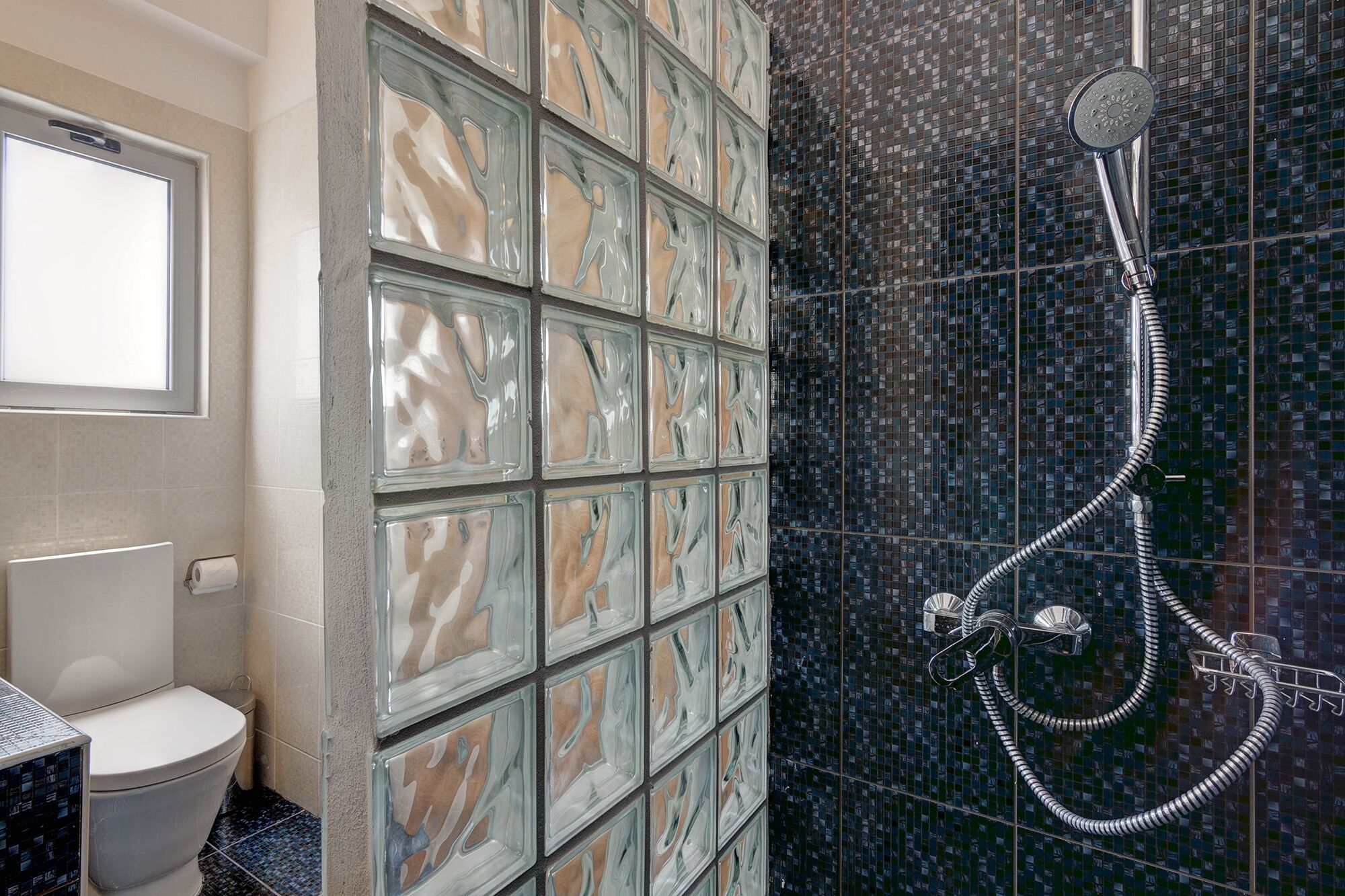 Une salle de bain avec des toilettes, une douche avec un pommeau flexible et un mur recouvert de carreaux décoratifs.