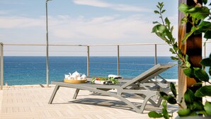 En terrasse med to liggestole, et bord med serveringsbakke med mad og drikke, og udsigt til havet.