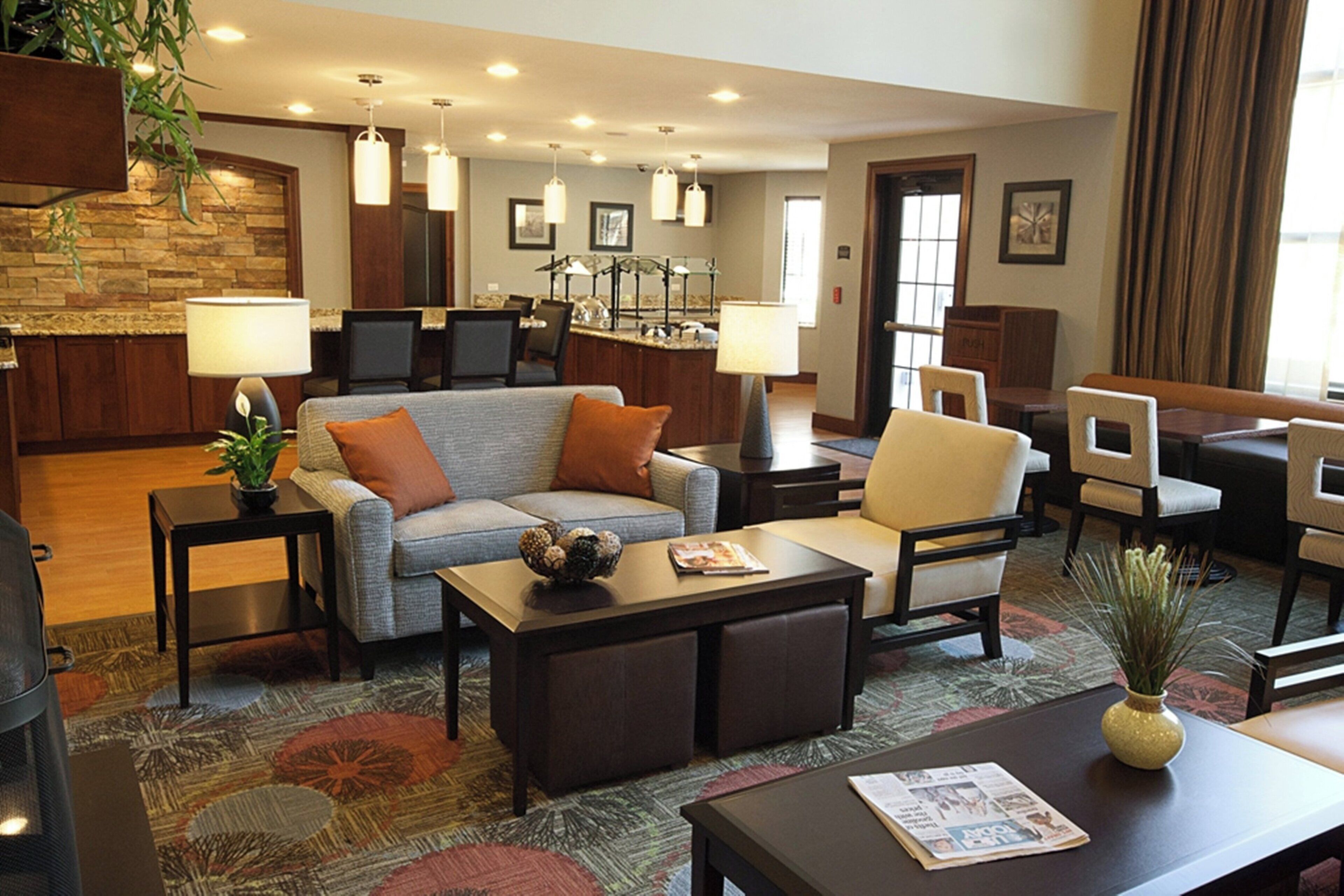 A hotel lobby with a sofa, armchairs, a coffee table, a fireplace, a kitchen area, and a dining area.