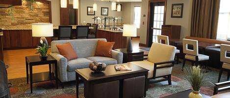 A hotel lobby with a sofa, armchairs, a coffee table, a fireplace, a kitchen area, and a dining area.