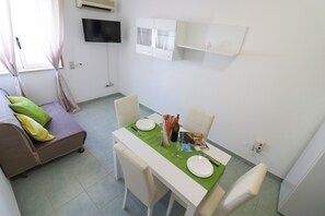 Un coin repas avec une table dressée pour deux personnes, un canapé avec des coussins, un téléviseur à écran plat fixé au mur et une fenêtre avec des rideaux.
