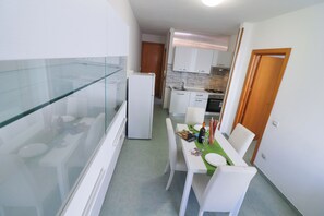 Une cuisine compacte avec des meubles blancs, une cloison en verre et un coin repas agrémenté d’une nappe verte.
