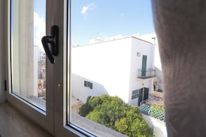 Et vindue med dørhåndtag, udsigt til en hvid bygning med balkon, og en grøn busk.