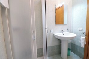 Une salle de bain avec une porte de douche en verre, un lavabo avec un miroir au-dessus et une poubelle verte.