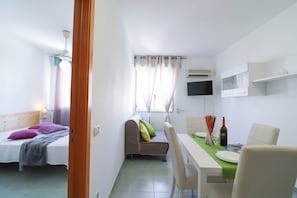 Une pièce de taille modeste comprenant un lit, un canapé, une table à manger avec des chaises, un téléviseur fixé au mur et un appareil de climatisation.