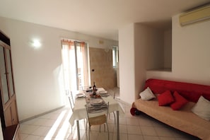 A dining area with a table set for two, a sofa with red cushions, and a tiled floor.