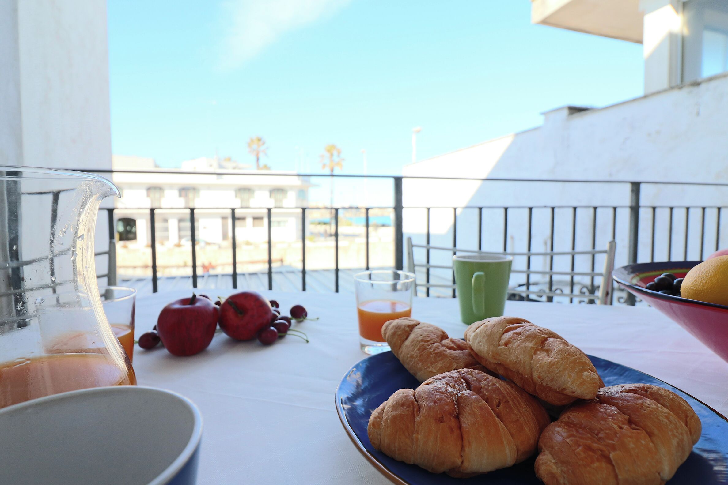 A balcony with a table set for breakfast, including croissants, a bowl of fruit, and a glass of orange juice.