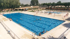 A large outdoor swimming pool with multiple lanes, surrounded by a fence and shaded by trees. - Camping Regio (Santa Marta de Tormes)