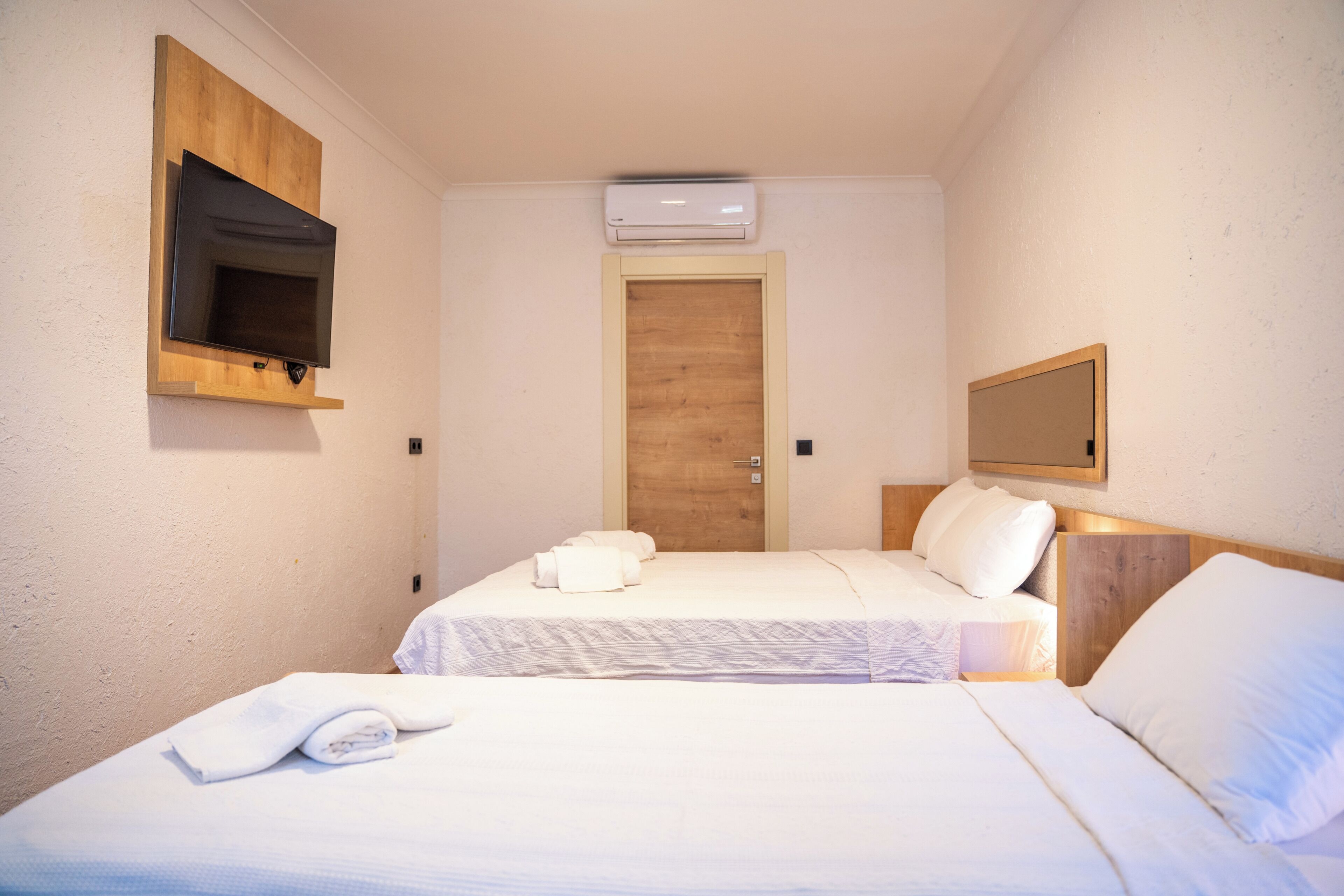 A hotel room with two beds, a TV mounted on the wall, an air conditioning unit, and a wooden door.