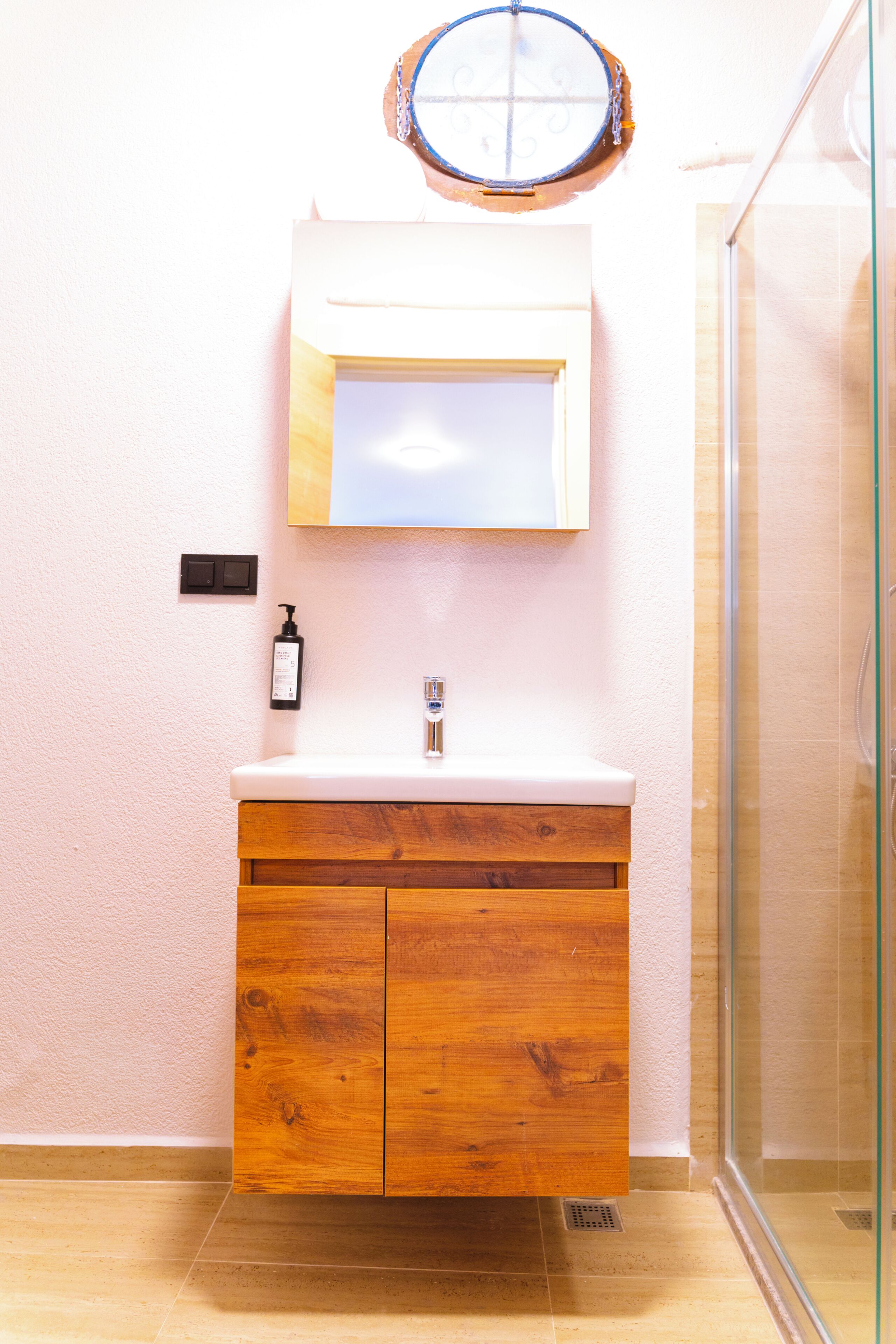 A bathroom with a wooden vanity, a mirror, a sink, a shower, and a wall-mounted light fixture.