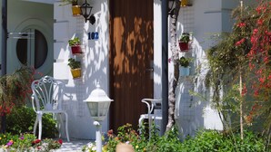 Un edificio blanco con puerta de madera, plantas en macetas, una silla blanca y una fuente de piedra.