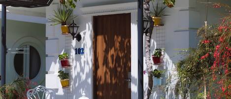 A white building with a wooden door, potted plants, a white chair, and a stone fountain.