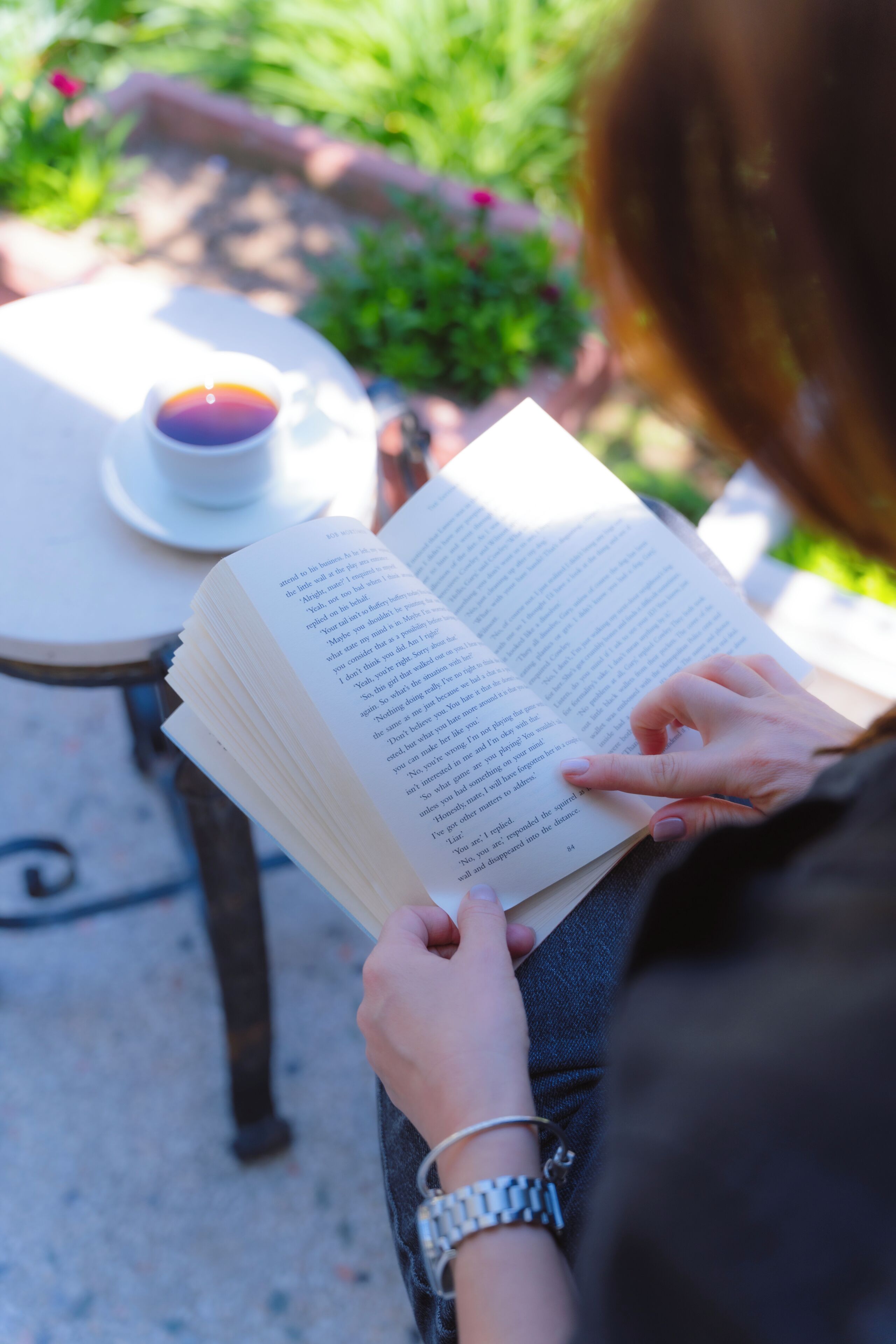 A person reading a book at an outdoor table with a cup and saucer.