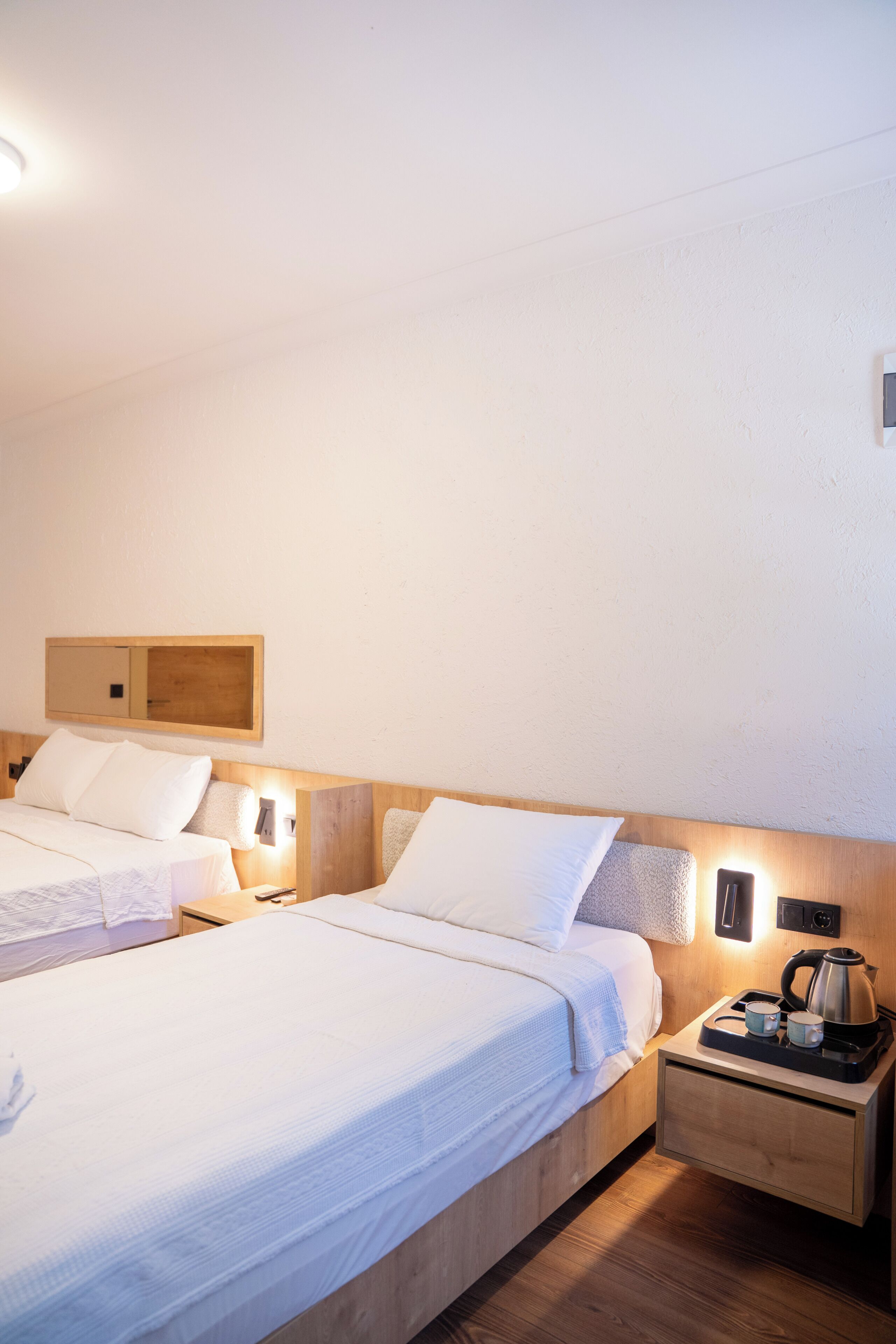 Una habitación de hotel con dos camas, cabecero de madera, lámparas de noche, mesita con tetera y tazas, y un espejo pequeño.