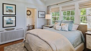 Un dormitorio con una cama grande, una mesita de noche, un velador, una ventana con vistas a zonas verdes y cuadros enmarcados en la pared.