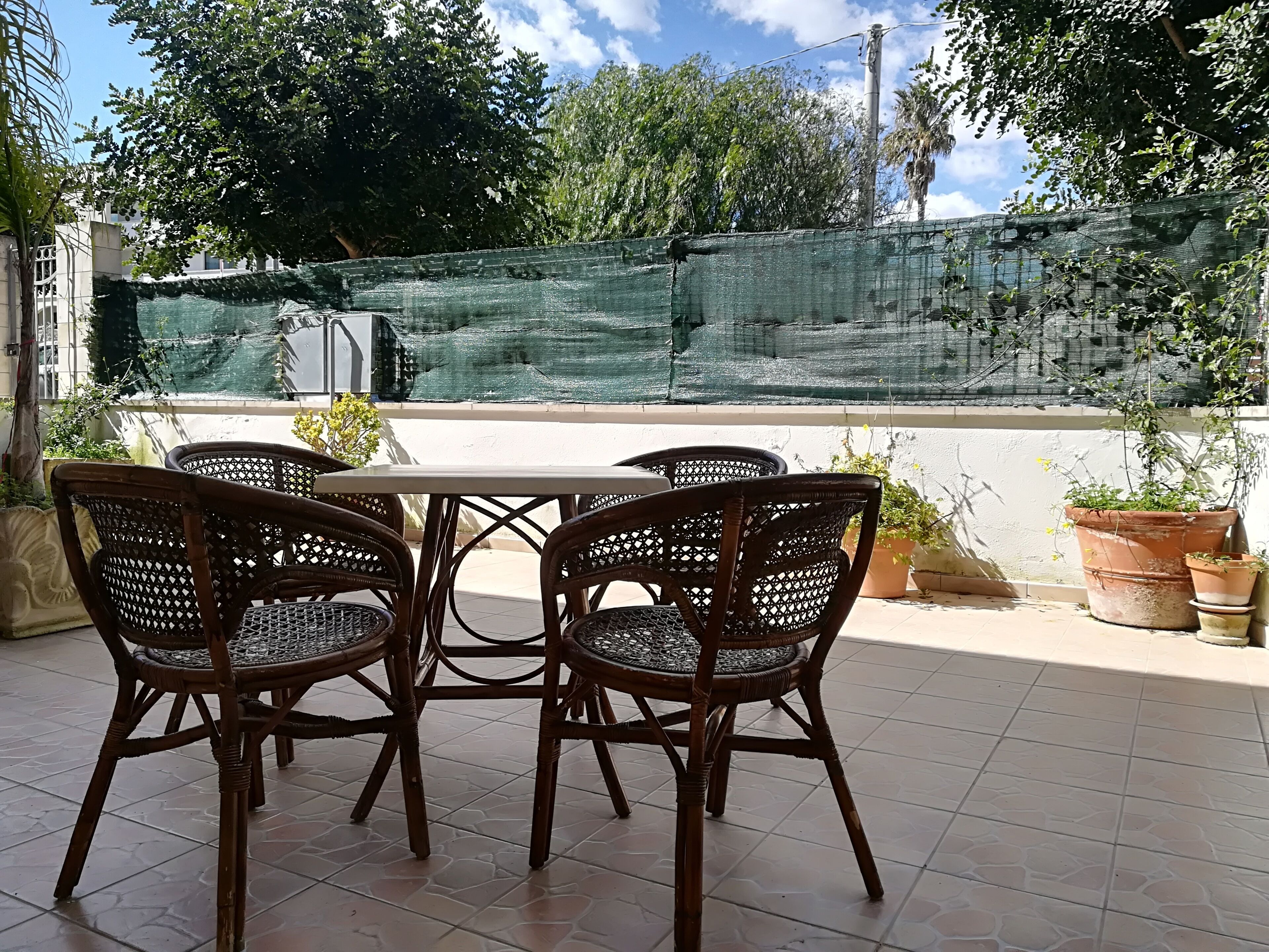An outdoor patio with a table and four wicker chairs, surrounded by potted plants and a green mesh fence.